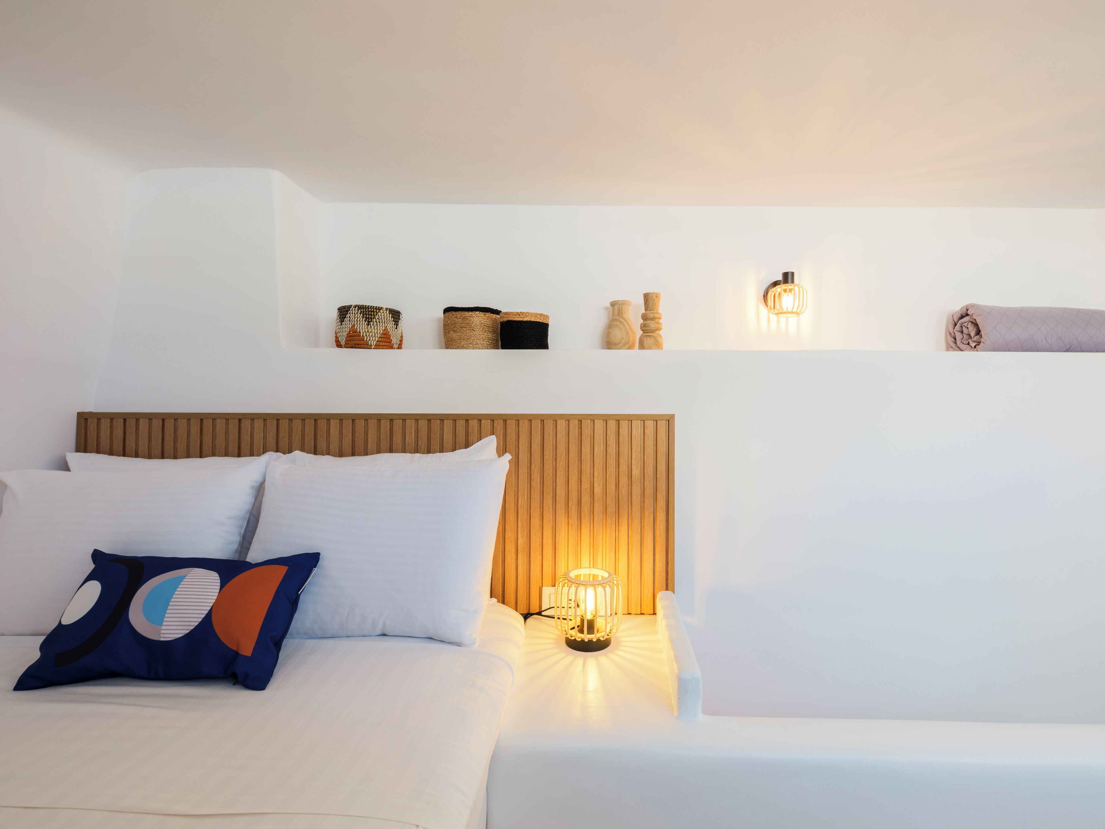 A neatly made bed with white linens and a wooden headboard, a bedside table with a lamp, and decorative items on a shelf above the bed.