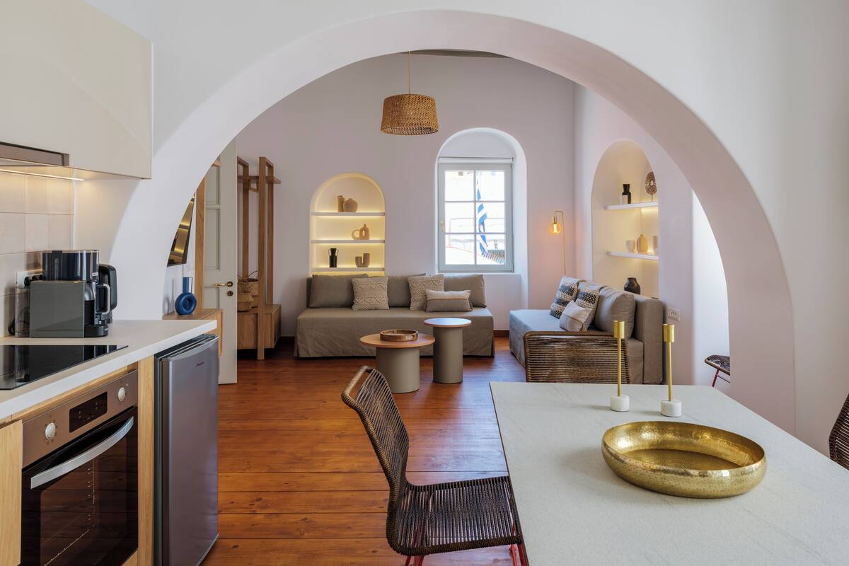 Una cocina moderna con horno integrado y cafetera, un comedor con mesa redonda y sillas, y una sala de estar con sofá y mesa baja.