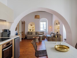 Uma cozinha moderna com forno embutido e máquina de café, uma área de refeições com mesa redonda e cadeiras, e uma sala de estar com sofá e mesa de centro.