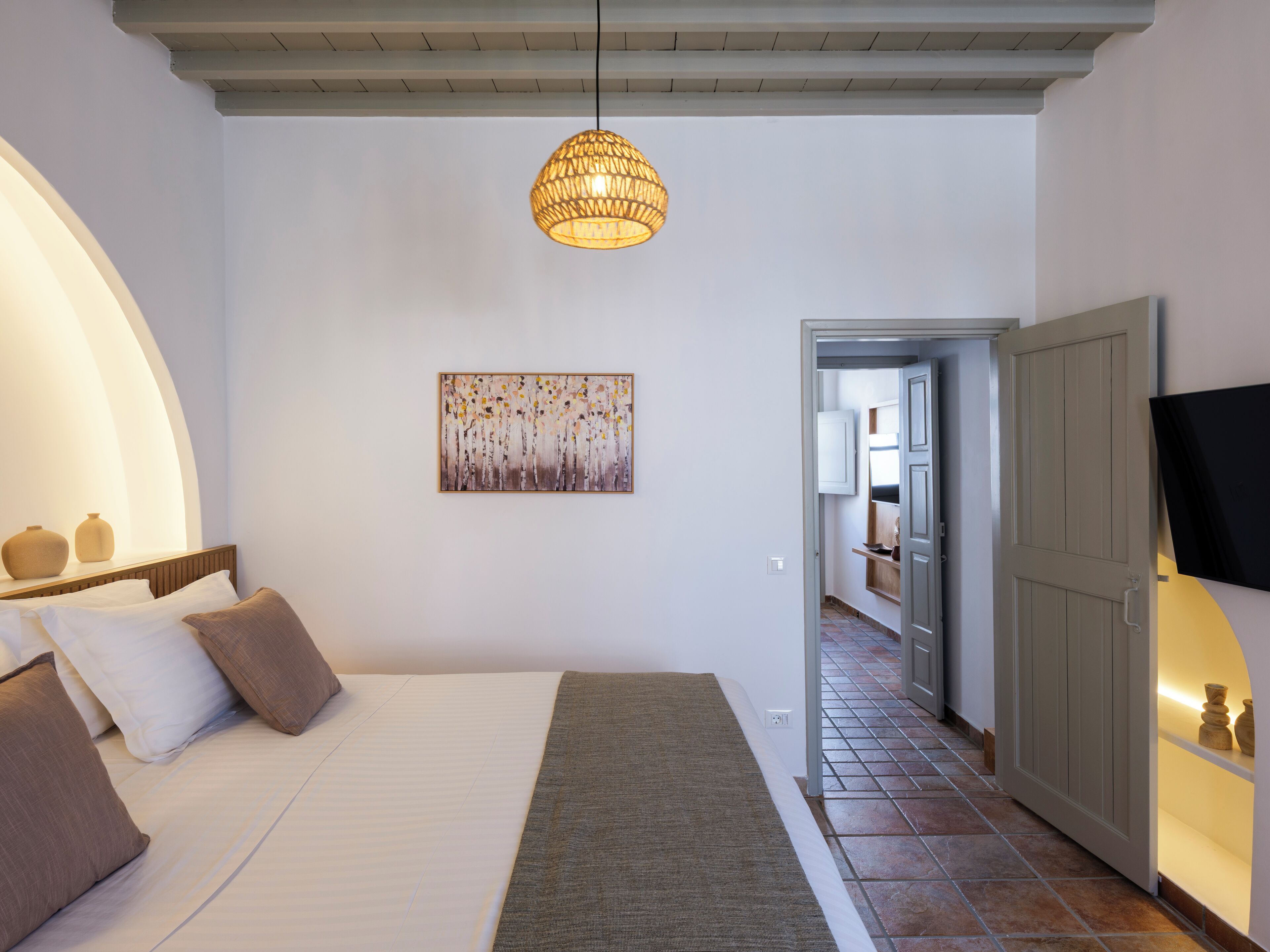 A bedroom with a bed, a painting, a hanging light fixture, and a door leading to another room.