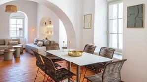 Un salón moderno con zona de comedor, que incluye una mesa blanca, sillas de mimbre y un sofá con cojines.