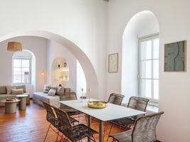 Uma sala de estar moderna com área de jantar, apresentando uma mesa branca, cadeiras de vime e um sofá com almofadas.