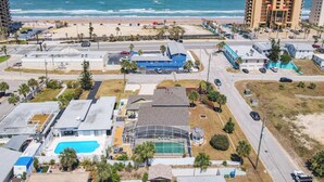 Vista aérea de una zona costera con playa, estacionamiento y edificios, incluyendo un hotel con piscina y un edificio azul.