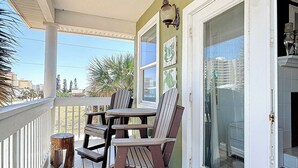 Un porche cubierto con una mesa y dos sillas Adirondack, una lámpara de pared decorativa y un puerta de vidrio que da al interior.