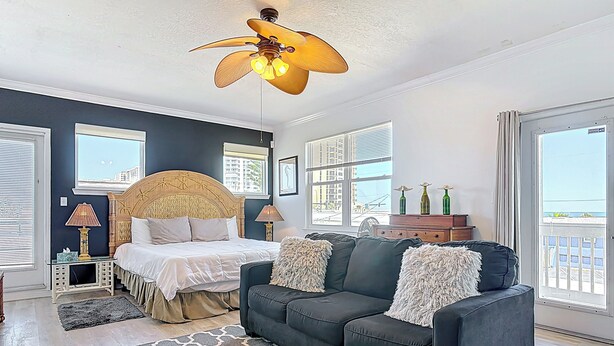 Un dormitorio con cama, sofá oscuro, alfombra estampada, ventilador de techo y ventanas grandes.