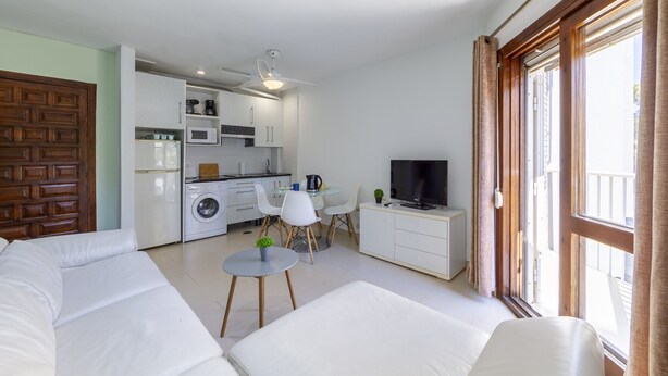 A modern living room with a sofa, a small table, and a TV. There is a kitchen area with a microwave, a coffee maker, and a washing machine. The room has large windows with curtains.