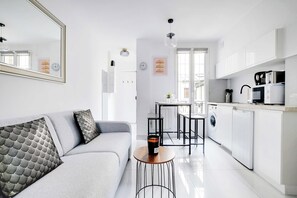 A modern kitchen with white cabinets, a microwave, and a coffee machine. There is a dining area with a table and chairs, and a sofa with patterned cushions. - Cosy, Fully-equipped apt - 4P - Montmartre (Paris)
