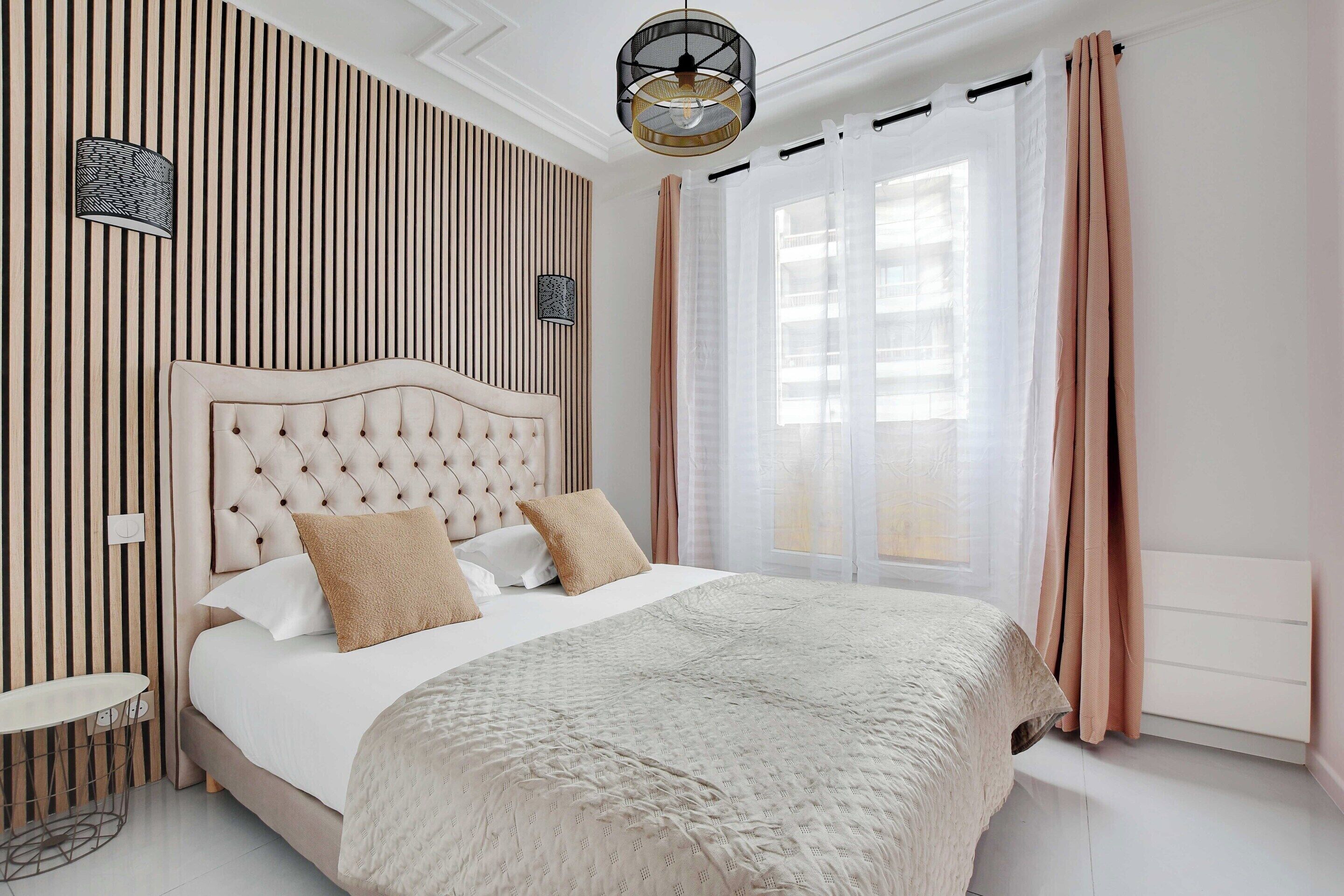 A bedroom with a tufted headboard, a large bed with white and beige bedding, a bedside table, and a wall-mounted lamp.