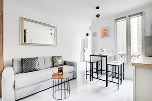 A modern living room with a sofa, a coffee table, a dining area with stools, a kitchen with a fridge, and a large window. - Cosy, Fully-equipped apt - 4P - Montmartre (Paris)