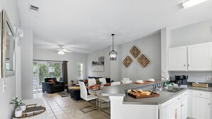 A modern kitchen with white cabinets, a central island, and a dining area with white chairs. - Disney Charm, Steps From Pool and Clubhouse! (Kissimmee)