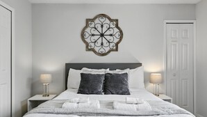 A neatly made bed with white linens, two bedside lamps, and a decorative wall piece above the bed. - Disney Charm, Steps From Pool and Clubhouse! (Kissimmee)