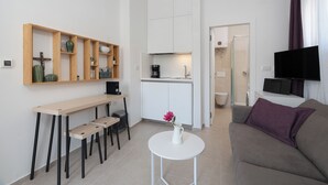 Un salon moderne comprenant un canapé, une petite table ronde, un coin cuisine avec des placards, une machine à café et un téléviseur fixé au mur.