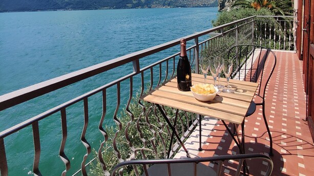 A balcony with a table, a bottle, and glasses, overlooking a lake with mountains in the background. - Peaceful Lakefront (Vello)