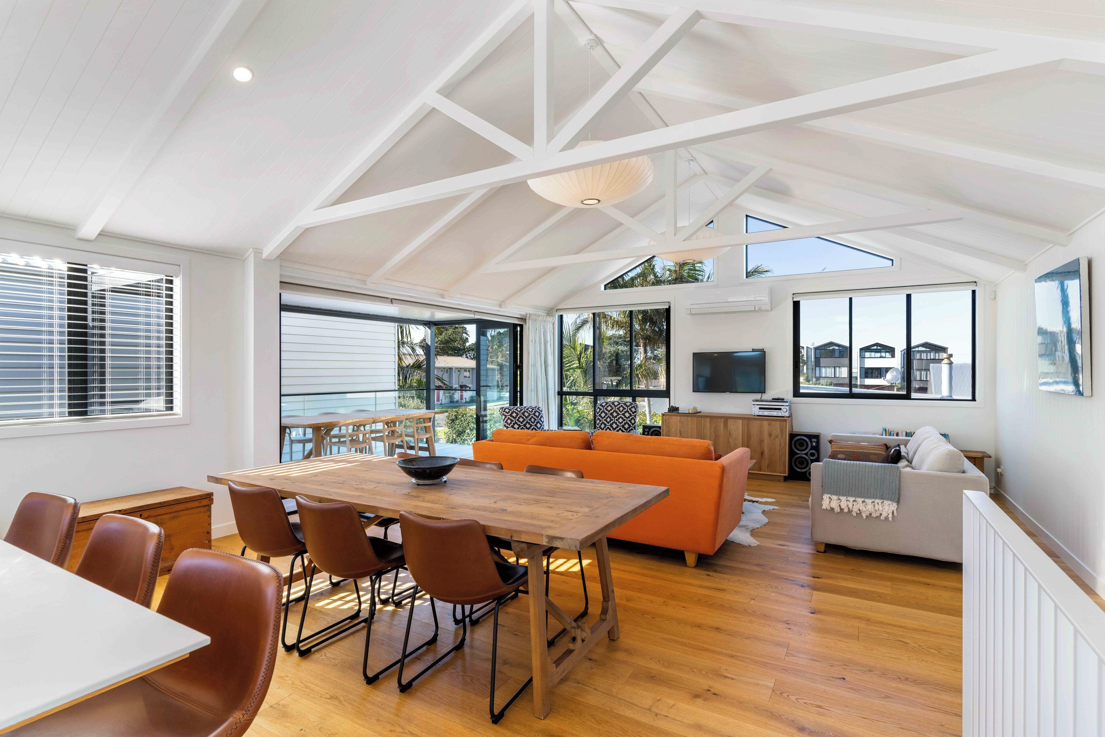 A spacious living area with a wooden dining table, orange sofa, and a TV mounted on the wall.
