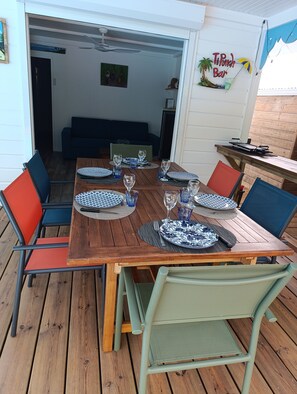 Uma área de refeições ao ar livre com mesa de madeira posta para quatro pessoas, rodeada por cadeiras coloridas.