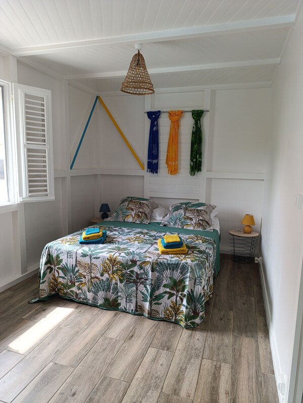 A bedroom with a bed, a hanging light fixture, a small table with a lamp, and a window with blinds.