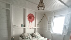 Un dormitorio con cama, lámpara colgante, ventana con contraventanas y una mesita de noche.