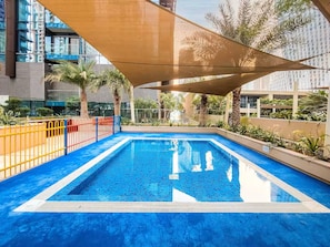 Una piscina rectangular con superficie de azulejos azules, rodeada por una valla amarilla y protegida por una gran toldo.