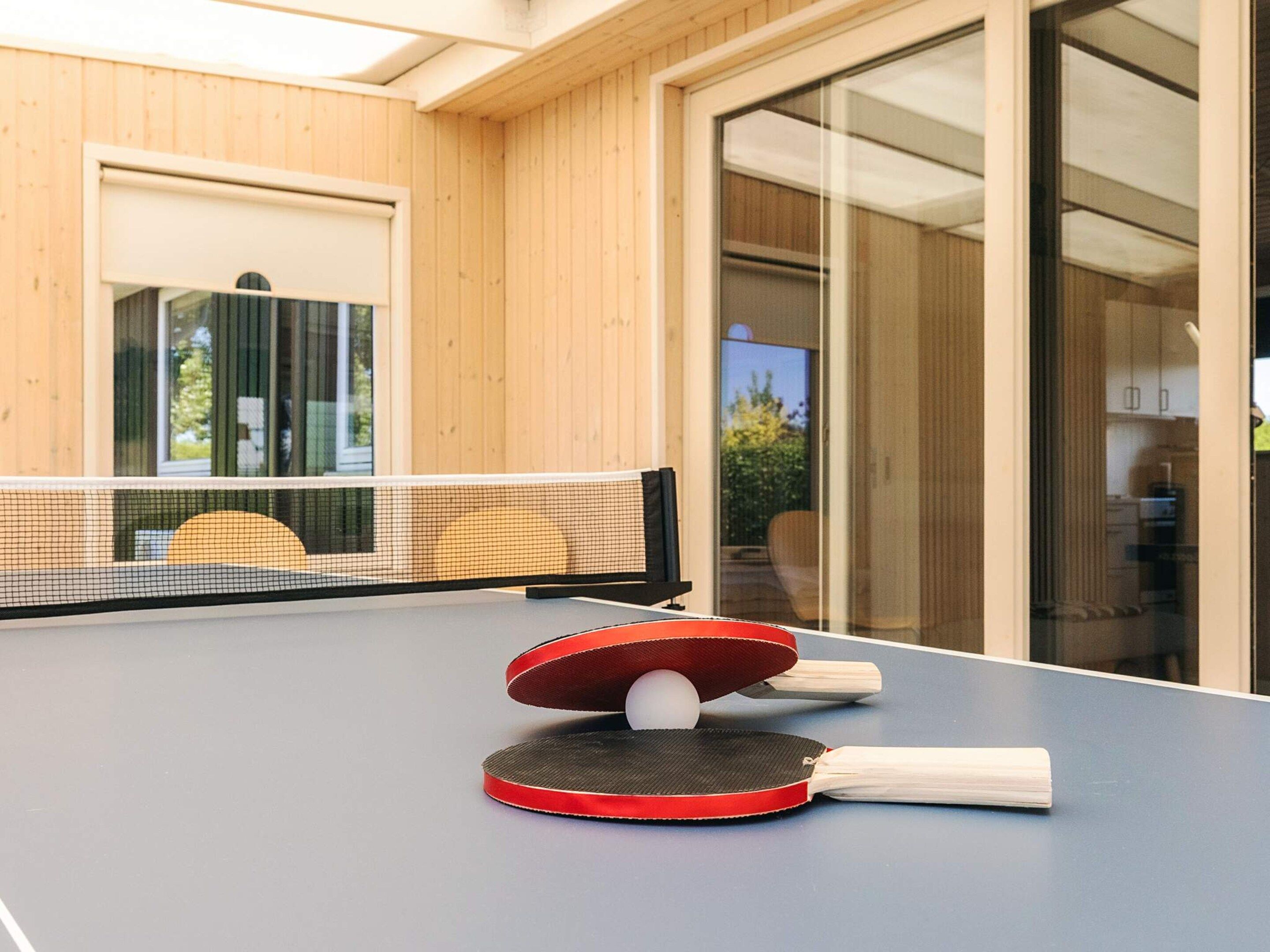 Ein Tischtennis-Tisch mit zwei Schlägern und einem Ball in einem Raum mit Holzwänden und einer Glastür.