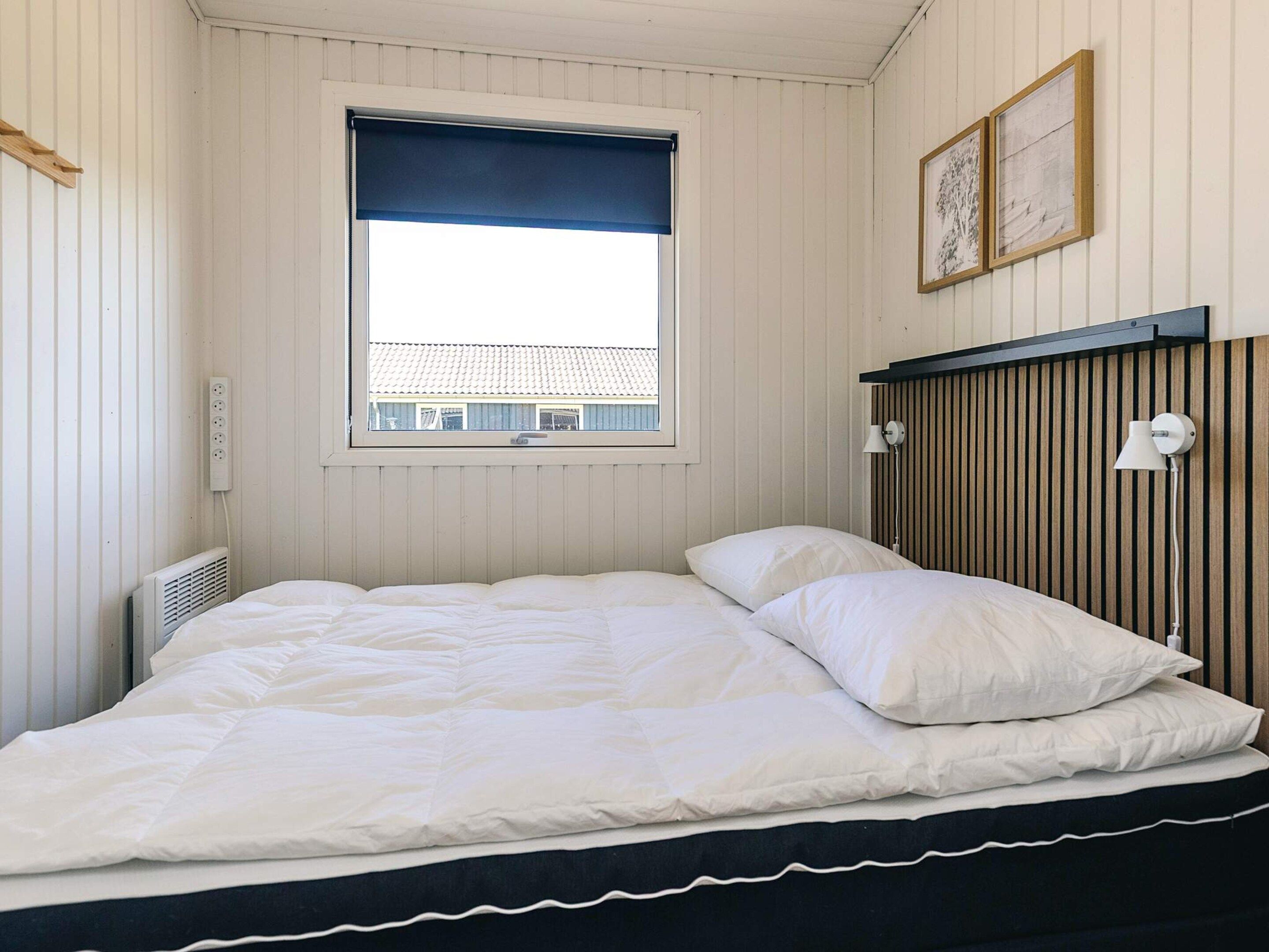 A single bed with white bedding, a window with a blue blind, a wall-mounted lamp, and a power outlet.