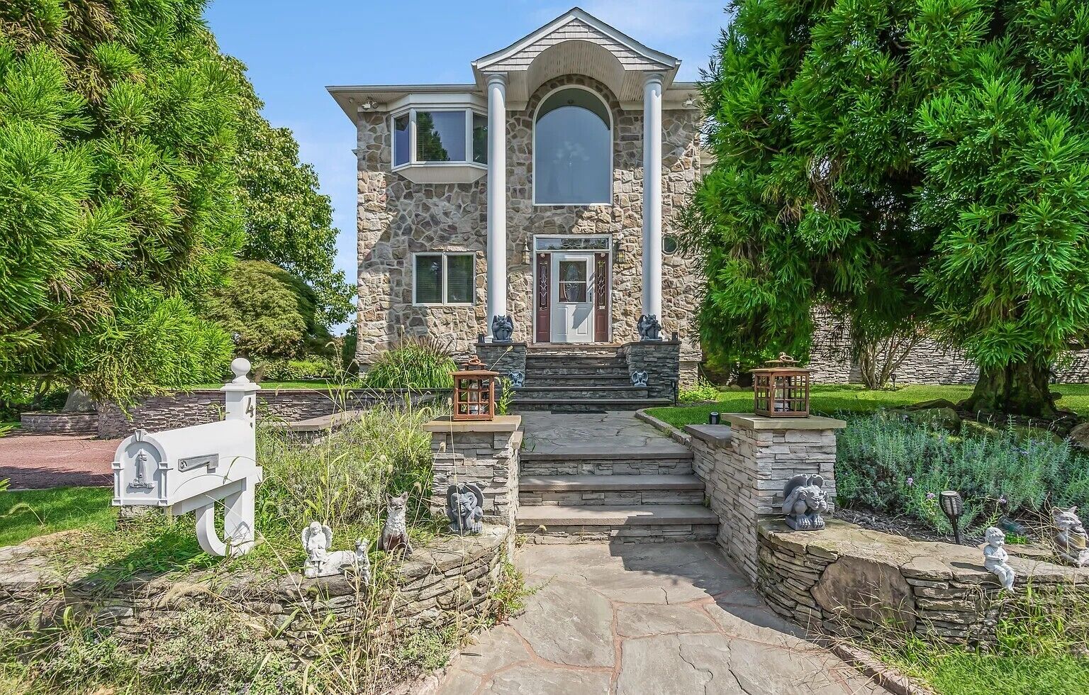 Une maison en pierre avec une entrée centrale, une boîte aux lettres et des statues dans le jardin.