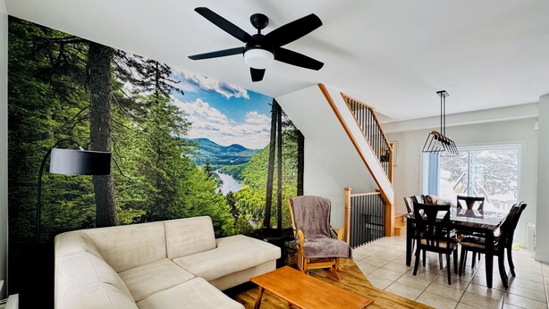 Un salon comprenant un canapé, une table basse en bois, un fauteuil, une table à manger avec des chaises, un ventilateur de plafond et une fresque murale représentant une forêt et des montagnes.