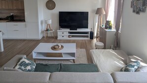 A modern living room with a flat-screen TV, a white sofa, a coffee table, and a kitchen area in the background.