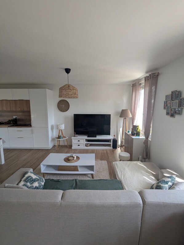 Sala moderna com televisão de tela plana, sofá branco, mesa de centro e área de cozinha ao fundo.