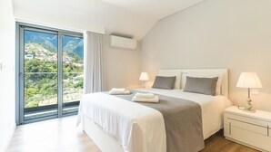 A bedroom with a large bed, a nightstand, a lamp, and a view of a mountainous landscape through a sliding door. - Villa Lorenzo by Atlantic Holiday (São Vicente)