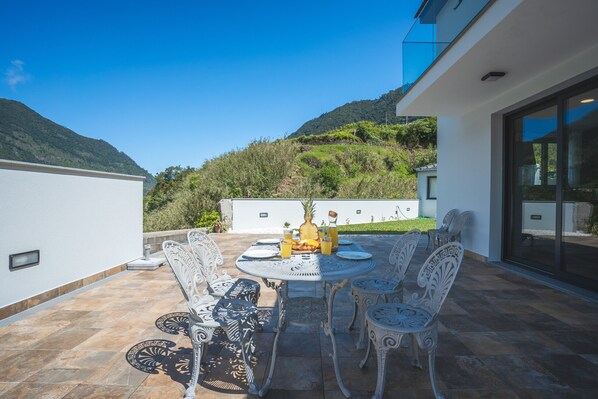Une terrasse extérieure avec une table ronde et des chaises blanches, entourée d’un mur blanc et offrant une vue sur une colline.