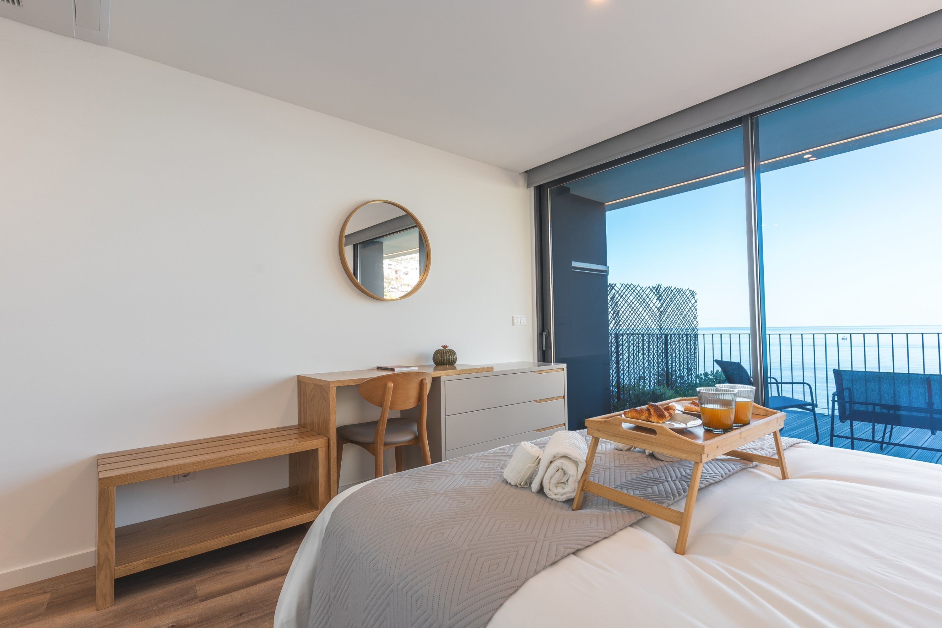 A modern hotel room with a large bed, a wooden desk, a chair, a small table with breakfast items, and a balcony with a view.