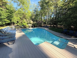 Uma piscina com deck de madeira, espreguiçadeiras e árvores ao redor.