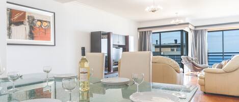 A dining area with a glass table set for four, a bottle of white wine, and a painting on the wall.