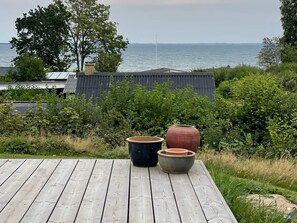 Eine Holzterrasse mit drei großen Pflanzgefäßen, Blick aufs Meer und einem Gebäude im Hintergrund.