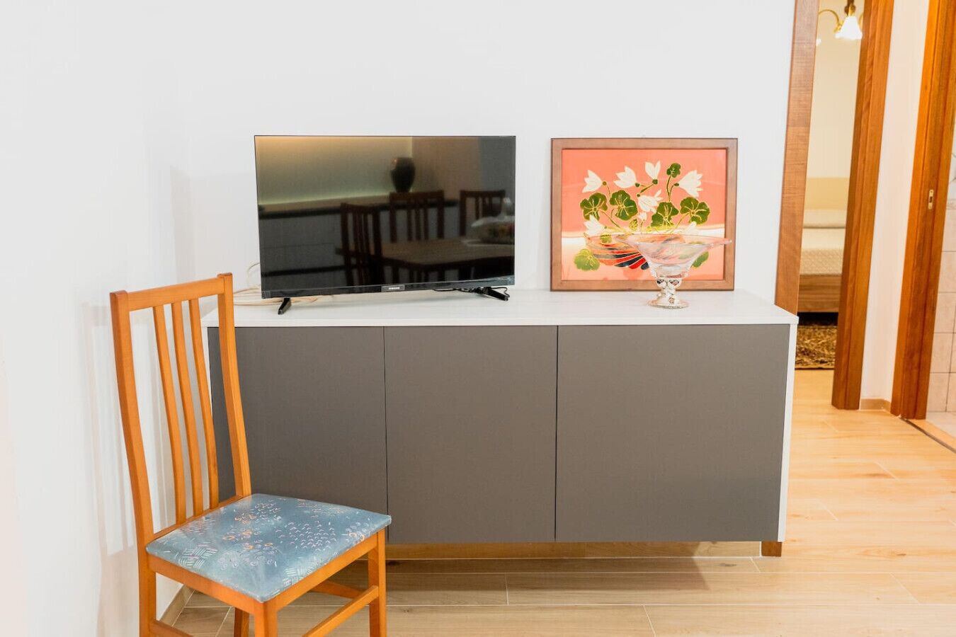 A modern living room with a grey cabinet, a wooden chair, a flat-screen TV, and a framed artwork on the wall.