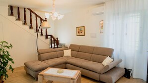 Una sala de estar con un sofá seccional marrón, una mesa de centro de madera, una escalera con balaustres de madera y una araña.