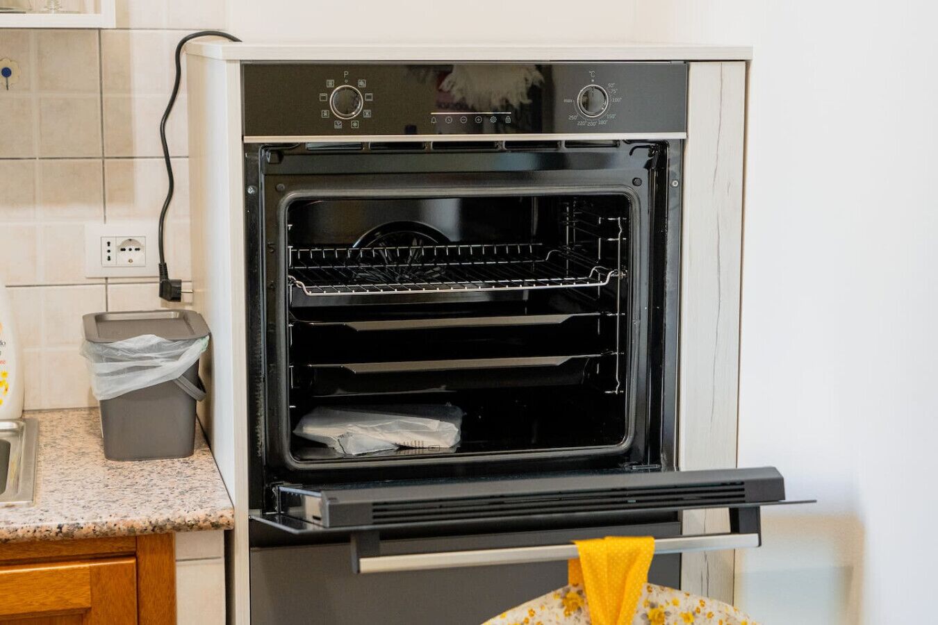 Ein offener Backofen mit Rost und einer Tasche darin, ein Mülleimer und eine Arbeitsplatte mit einem Wasserhahn.