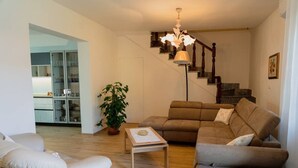 Una sala de estar con un sofá, un sillón y una mesa de centro. Se observa una escalera con una araña.
