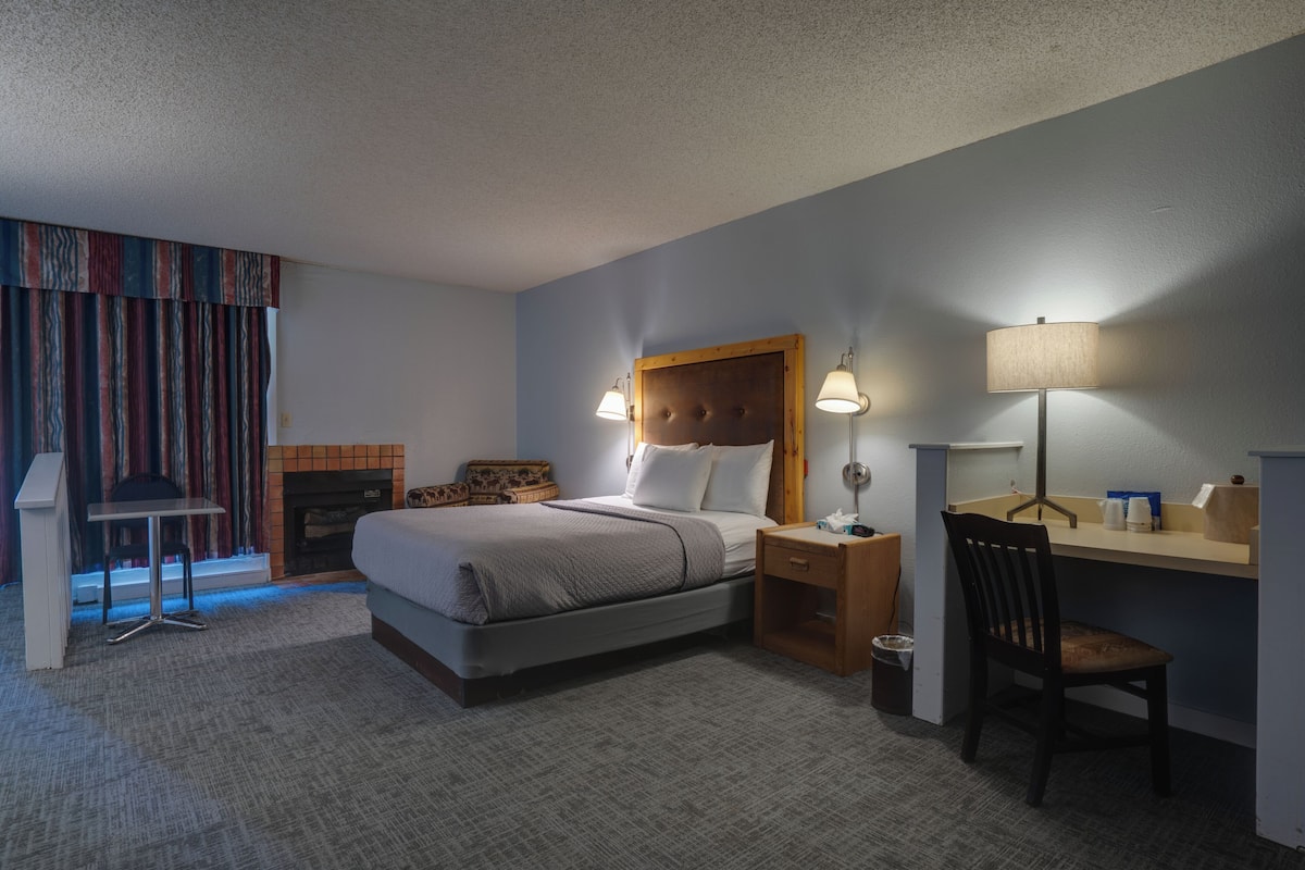 A hotel room with a bed, bedside lamps, a desk, a chair, a small table, and a fireplace.