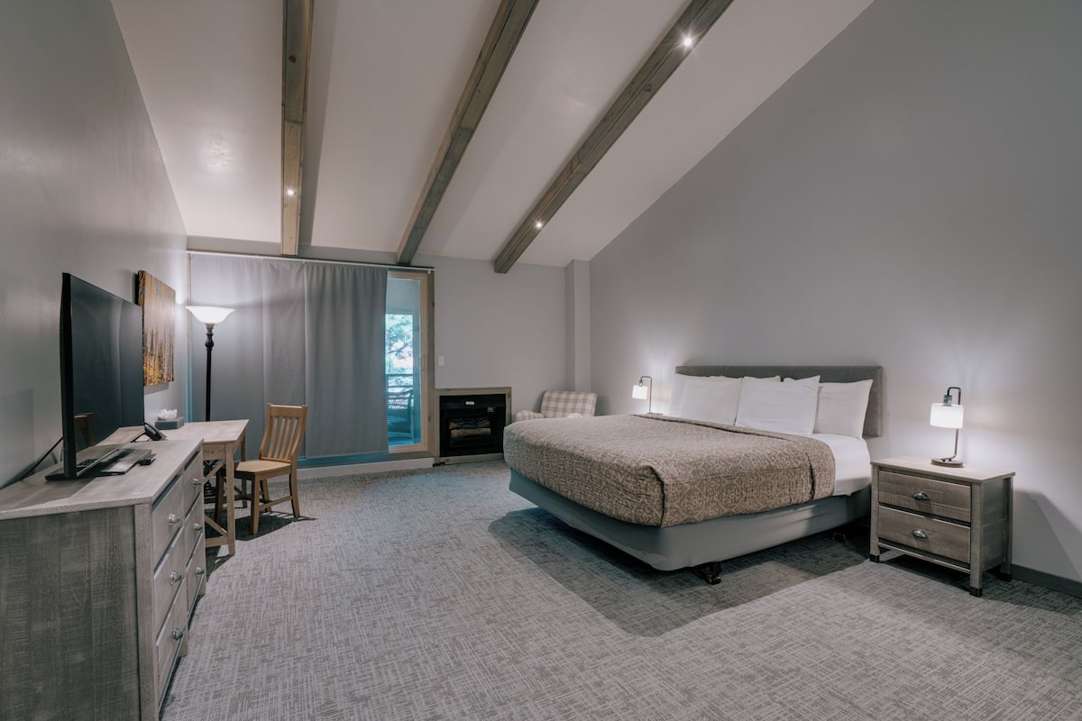 A modern bedroom with a bed, bedside tables, a desk, a chair, a TV, and a fireplace.