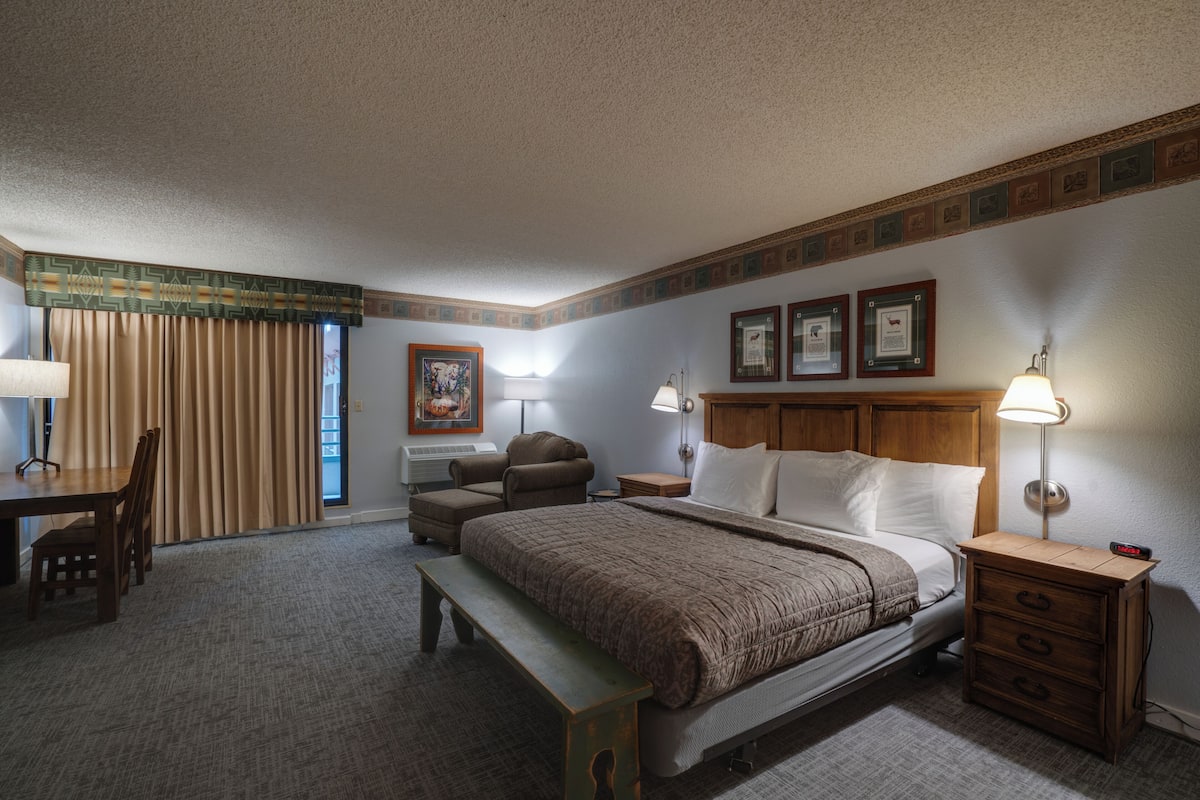 A hotel room with a bed, bedside tables, a desk, a chair, a bench, and a window with curtains.