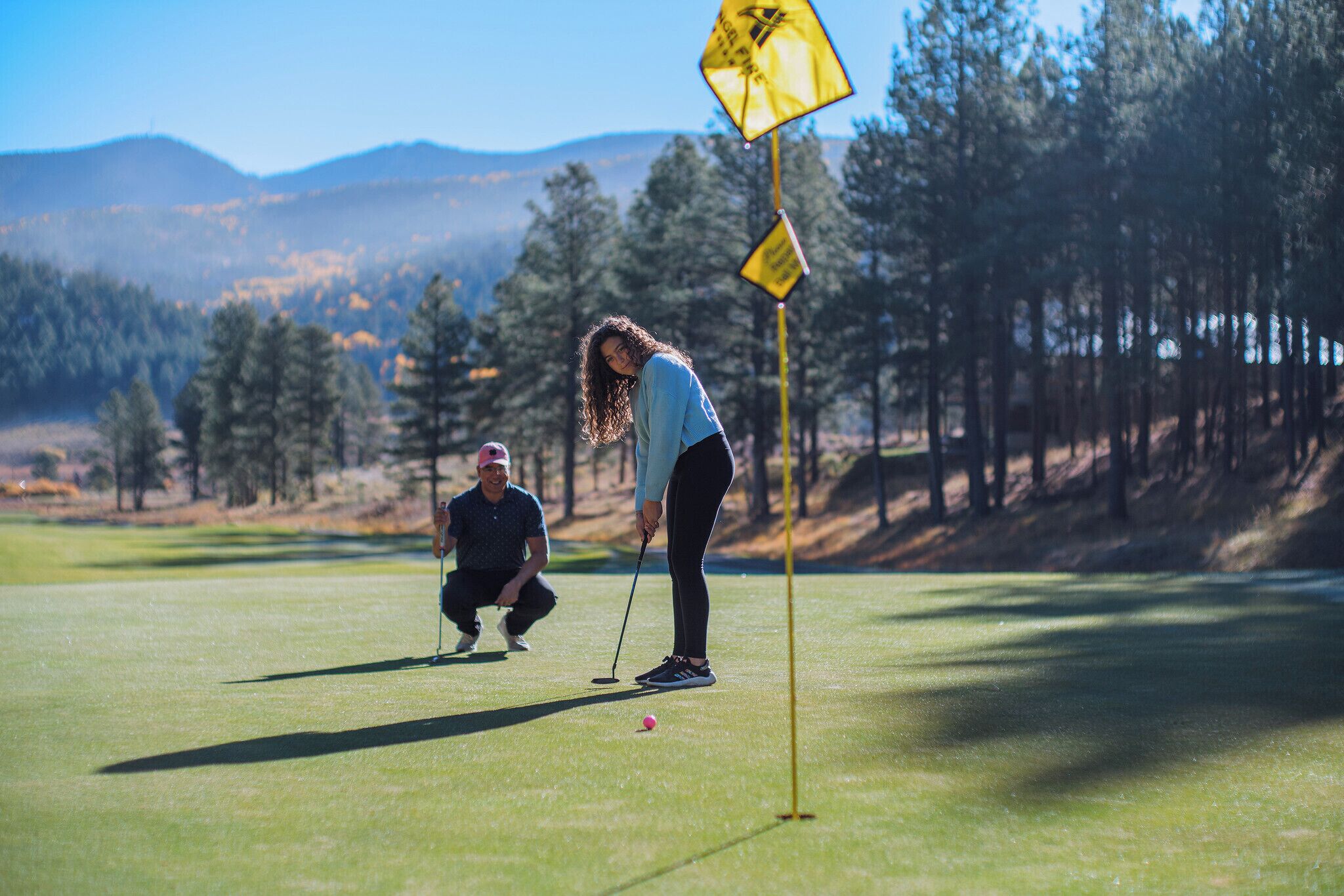 A golf course with a flagstick, a putting green, and a forested background.