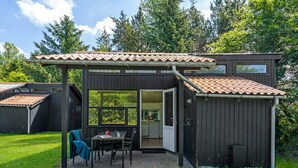 A compact, dark-colored wooden house with a tiled roof, a patio with a table and chairs, and a large window. - 4 Person Holiday Home in Hampen (Hampen)