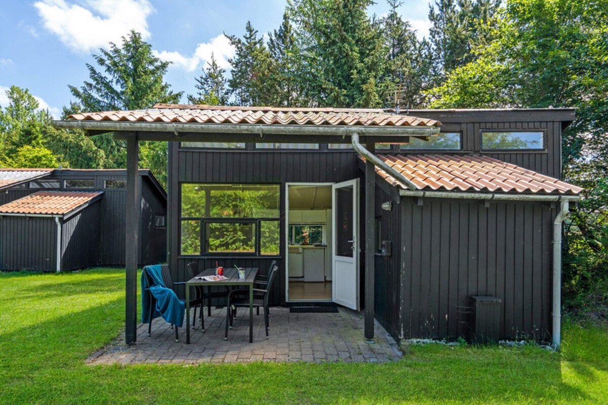 A compact, dark-colored wooden house with a tiled roof, a patio with a table and chairs, and a large window.