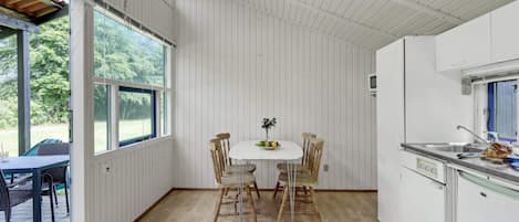 A room with a dining table, chairs, a kitchen area with a sink, and large windows allowing natural light.