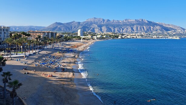 A coastal cityscape with a beach, clear blue sky, and mountains in the background.