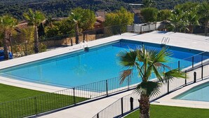 A rectangular swimming pool with a clear blue water, surrounded by a metal fence, and a view of the sea and hills in the background.
