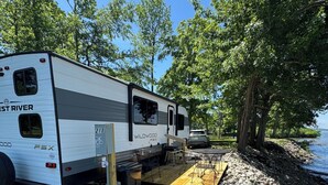 Een witte camper met 'WEST RIVER' en 'WILDWOOD'-logo's, geparkeerd aan een rotsachtige kust met een houten terras.