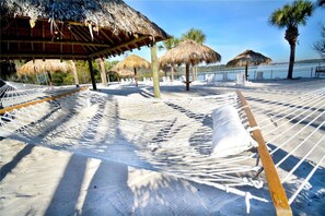 A hammock set up on a sandy beach with a thatched-roof structure in the background.