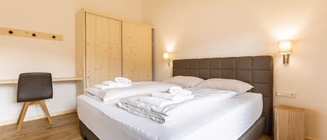 A neatly made bed with white linens and towels, a wooden wardrobe, a small bedside table, and a chair.